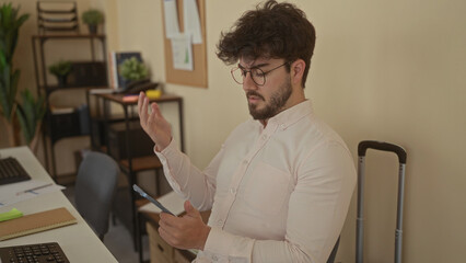Young man with beard using smartphone in modern office setting with indoor plants, bookshelves, and a suitcase.