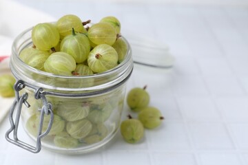Fresh green gooseberries in jar on white tiled table, closeup. Space for text