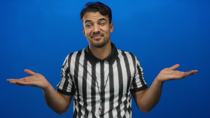 Fototapeta premium Young hispanic man referee in striped shirt gestures with uncertainty against an isolated blue background.