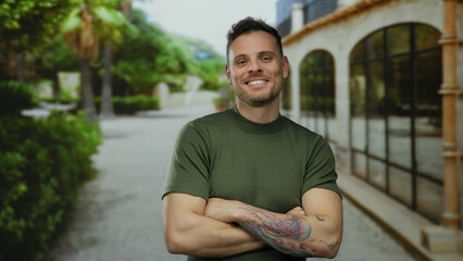 Fototapeta premium Smiling man with tattoos in a green shirt stands with crossed arms on a sunny urban street lined with trees and buildings creating a relaxed and confident atmosphere