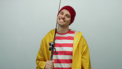 Fototapeta premium Young man holding a fishing rod, wearing a yellow raincoat and red beanie, smiling confidently against a white background, embodying a handsome, cheerful, and casual vibe.