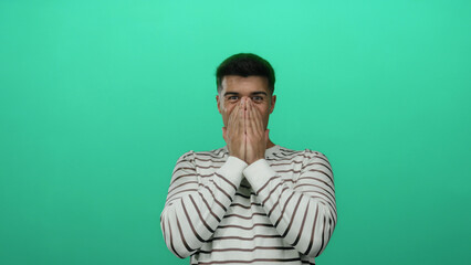 Fototapeta premium Young man covers mouth with hands joyfully standing against an isolated green background wearing a striped shirt showcasing emotion and surprise.