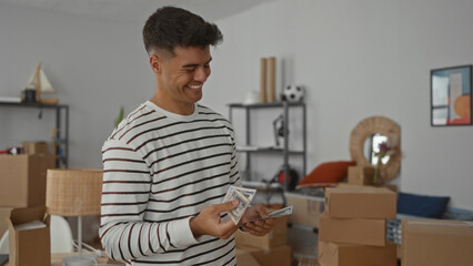 Fototapeta premium Young man in striped sweater smiling and counting dollar bills in living room surrounded by moving boxes, reflecting new home excitement and transition.