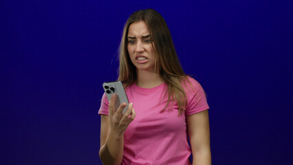 Woman expressing frustration while holding smartphone against blue background, wearing pink shirt,...