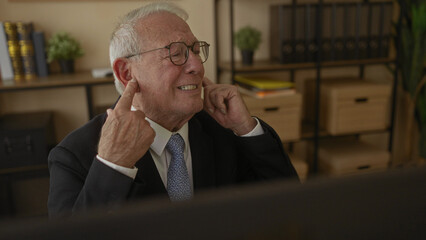 Senior man in a suit sits in an office covering ears with fingers, expressing frustration indoors...