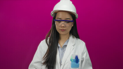 Young chinese woman architect studies blueprints wearing hardhat against isolated pink background, focusing intently on construction plans with a vibrant wall enhancing the scene.