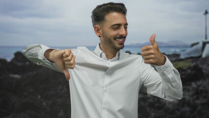 Man displays thumbs up and thumbs down gestures in white shirt at rocky seaside beach while smiling broadly  conflicted. © Krakenimages.com