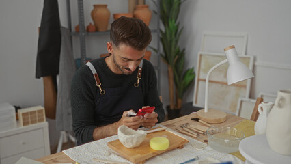 Man in apron holds red smartphone tapping screen beside clay bowl on wooden table with pottery tools in workshop; focused creativity.
