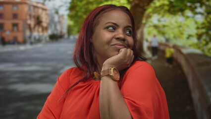 Fototapeta premium Woman in orange blouse resting chin on hand on sunlit city street under leafy trees; quiet contemplation.