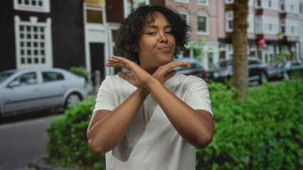 Young african american woman crosses bare arms in refusal gesture on busy city street near parked car and leafy hedge; rejection.