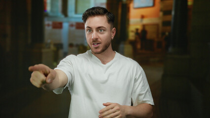 Young man with a beard stands on a city street, expressing surprise, wearing a casual white shirt in an urban outdoor environment at night, hands covering mouth.