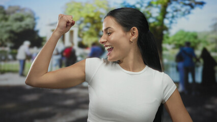 Young woman in a park flexing her arms with a joyful expression against a blurred outdoor background, exuding confidence and strength in a casual setting.