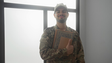 Man in camouflage uniform holds stack of books with one hand against window frame inside building; pride duty.