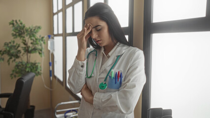 Young hispanic woman doctor in uniform with stethoscope looking stressed in a hospital clinic room, highlighting the challenges of her profession in a medical environment.