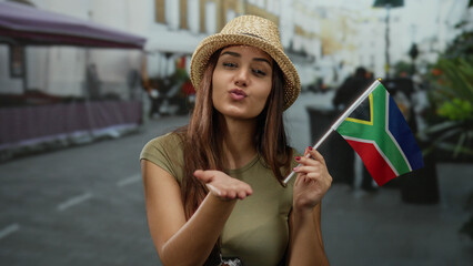 Woman smiling and blowing a kiss with south african flag on an outdoor street while wearing a straw...