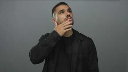 Young hispanic man with hand on chin and head tilted upward in grey studio showing pensive pose; idea reflection.