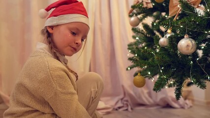 A slow motion closeup of a happy Caucasian girl decorating Christmas tree indoors at home. Young child wearing festive hat carefully hangs baubles on fir with joyful expressions. Holiday preparation - Powered by Adobe