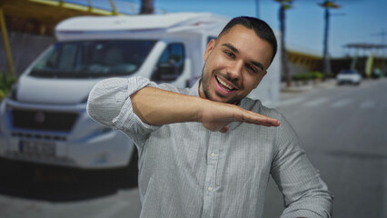 Man smiling with raised hand gesture in front of modern white camper van on a sunny city street;...