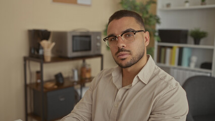 Young hispanic man wearing glasses glances to the side at a coffee station on a shelf in an office workspace; contemplation.