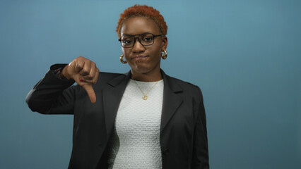 African american woman in black blazer and glasses showing thumb down with pursed lips in blue...