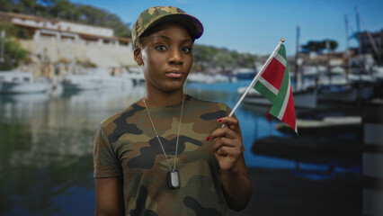 Young woman in camouflage uniform holds suriname flag aloft on street lined with boats and...