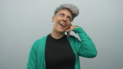 Woman wearing green cardigan laughs while mimicking a phone call gesture with finger to ear in...