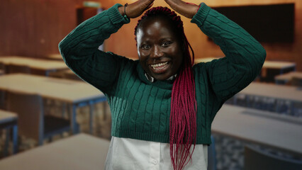 Woman gesturing playfully in a classroom setting with hands on head and vibrant pink braids under green sweater, expressing excitement or fun indoors.