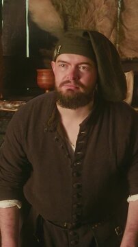 Bearded keeper in bonnet waits for guests in tavern. Stubby dwarf innkeeper stands by counter against rustic wooden shelves in store. Fantasy character