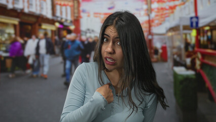 Hispanic woman wearing light blue top points finger to chest on bustling street with determined...