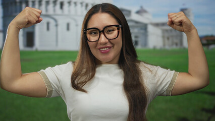 Young hispanic woman flexing bare arms in front of pisa tower building under clear sky; strength empowerment confidence.