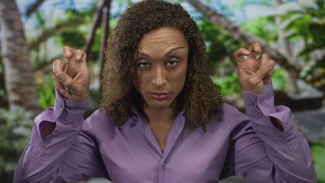 Transgender woman in purple shirt raising both hands to display peace signs amid green foliage in park; peace pride.
