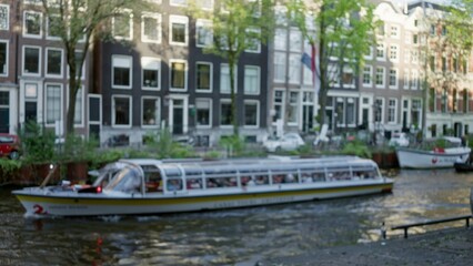 Blurred canal in amsterdam with a defocused tourist cruise boat passing historic buildings under green trees, capturing the serene ambiance of the netherlands.