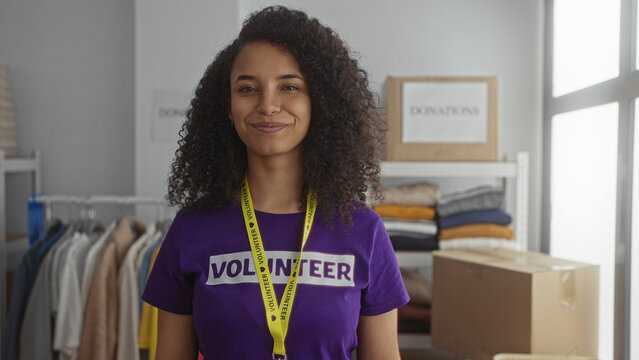 Young hispanic woman smiling in purple volunteer shirt at indoor charity center with clothing donations in the background. - Powered by Adobe