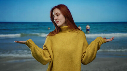 Woman with red hair and yellow sweater standing on beach with ocean in background, embodying peace and harmony amidst scenic seaside setting.