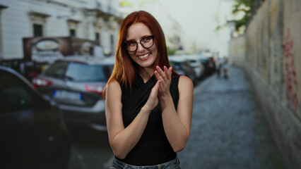 Woman smiling outdoors in a city street setting with cars parked along the side, exhibiting joy and confidence in an urban environment, showcasing casual fashion and red hair.