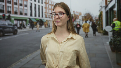 Obraz premium Young blonde woman wearing glasses standing and observing a city street lined with shops in broad daylight; alertness.