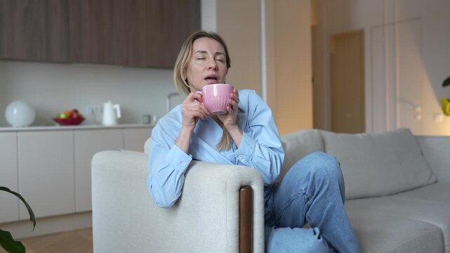 Relaxed interested middle aged woman drink hot tea or coffee taking mindful break with positive thoughts at home. Pleased mature female holding warm beverage sitting on cozy sofa enjoying aroma in mug