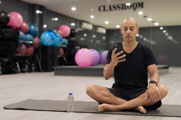 Fototapeta premium Hispanic bald man athlete in gym classroom doing yoga meditation while sitting and using phone