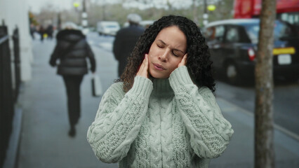 Young woman on street experiencing headache with eyes closed and pained expression surrounded by urban scenery and pedestrians.