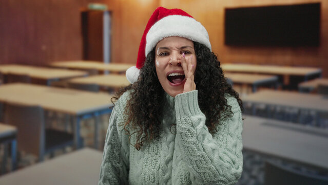 Woman in santa hat speaking joyfully in an empty classroom during holiday season, blending festive spirit with a learning environment.