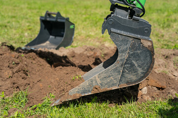 Excavator digging bucket breaking ground on grassy construction site with spare bucket in background for trenching, earthmoving, or landscaping projects