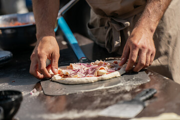 Artisan cook shaping pizza on peel, topping dough with fresh onion and ham slices, rustic outdoor food preparation before wood fired baking.