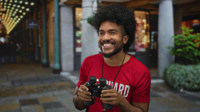 African american man in lifeguard shirt holding binoculars with a bright smile standing on a lively city street during night time.