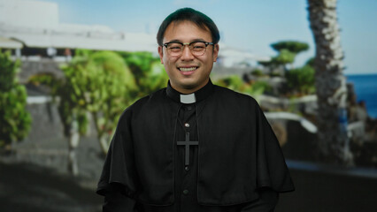 Asian man in priest attire smiles and gestures upward on a sunny seaside promenade with greenery and ocean in the background.