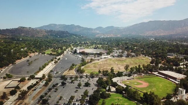 Baseball field, parking cars and rose bowl stadium with mountains in background. Sunny day summer day in Pasadena, California. Drone wide shot.