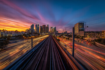 Sunset view from an elevated train track above a busy intersection, light trails from cars below and urban skyline in background