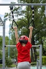Obraz premium Child in red shirt and helmet hanging on gym rings at outdoor playground.