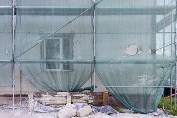 Building under renovation with green mesh construction netting and window behind scaffolding. Dusty facade, wooden barrier and debris on ground.