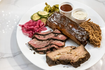 This plate showcases a barbecue meat assortment with beef brisket, pastrami, ribs, and pulled pork. Sauces accompany with pickles and pickled onions.