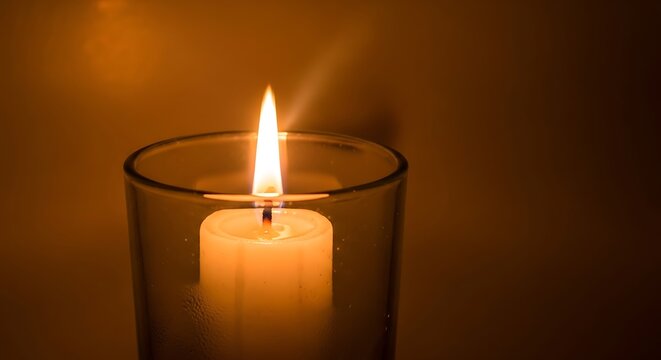 A single white candle burning brightly inside a clear glass holder, casting a warm, soft glow against a dark background.